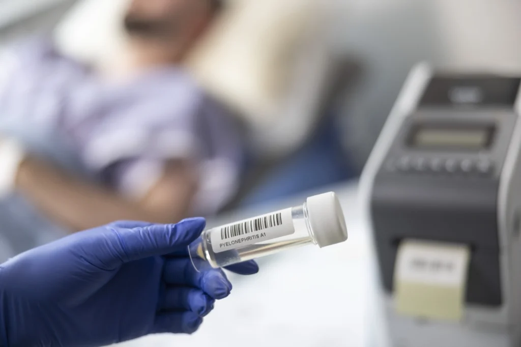 A gloved hand holds a barcode-labelled specimen tube beside a label printer at a patient bedside