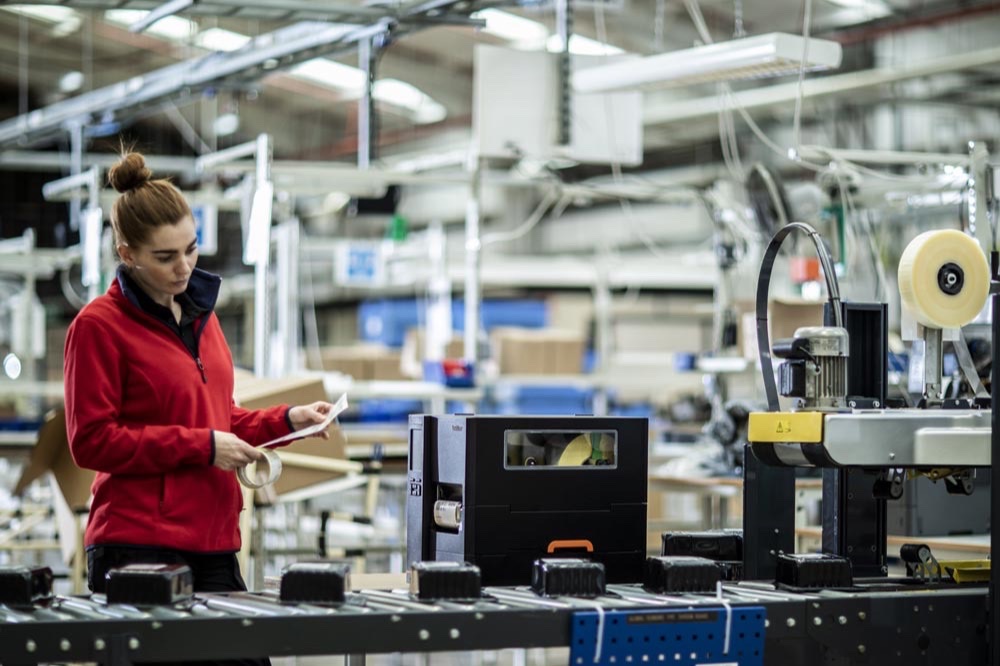 A production worker reads a printed label beside an industrial label printer on a factory line
