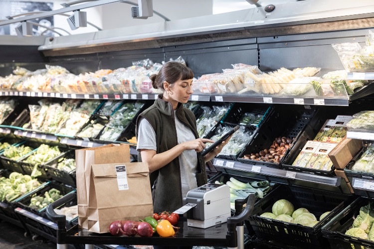 A shop assistant checks a tablet beside a chilled produce aisle with a label printer on the trolley