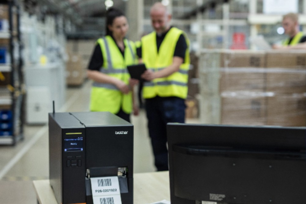 A Brother industrial label printer printing barcode labels at a dispatch bench, with depot staff checking a tablet behind it